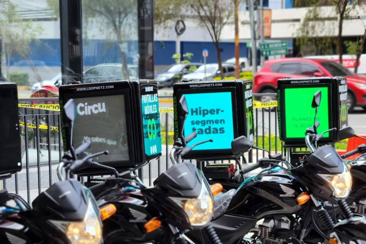 Varias motos de reparto aparcadas en fila en una calle de Ciudad de Guatemala. Cada una lleva una caja con pantallas LED encendidas mostrando mensajes de Loopnetz.