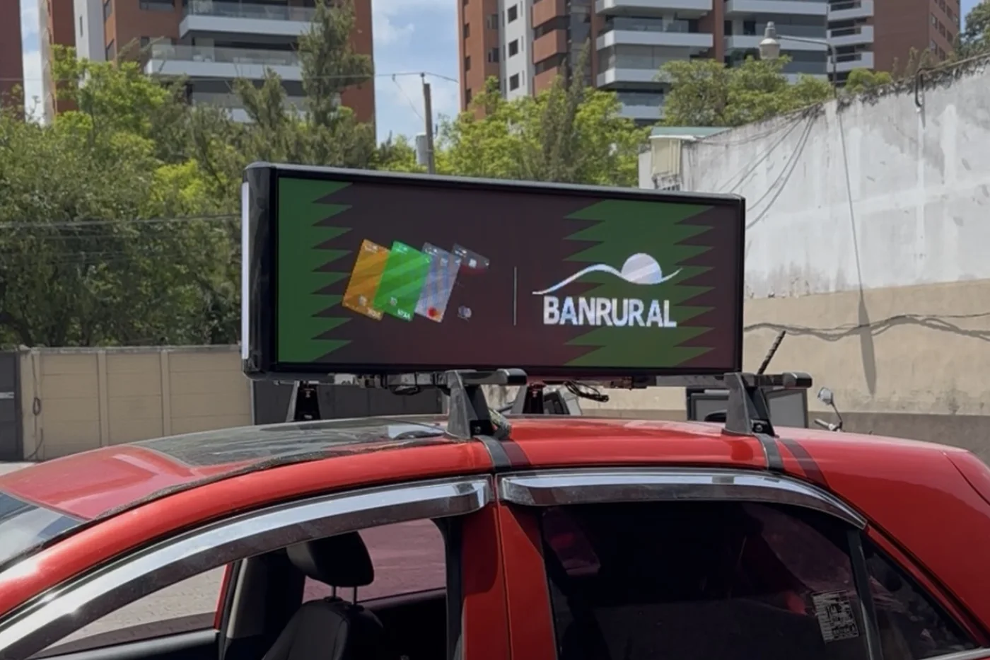 Rótulo LED de dos caras montado sobre el techo de un carro rojo en una avenida de Ciudad de Guatemala, con edificios al fondo. La pantalla muestra un anuncio de Banrural con tarjetas de crédito.