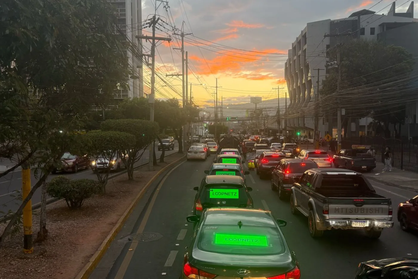 Avenida de Ciudad de Guatemala al atardecer, con el tráfico detenido. Dos vehículos delante llevan pantallas LED encendidas en la luneta trasera, con el mensaje de Loopnetz en verde.